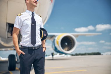 Neşeli erkek pilot havaalanında uçağın yanında duruyor.
