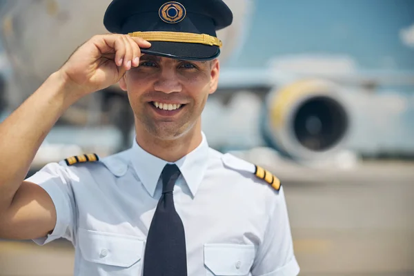 Handsome pilot in command touching captain hat and smiling — Stock Photo, Image