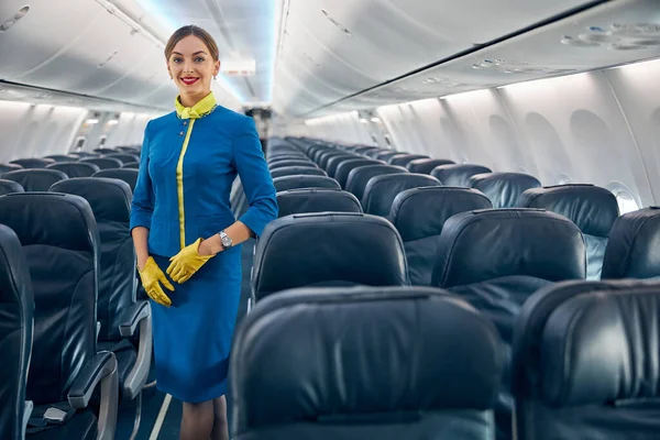 Молодая привлекательная женщина в синей форме позирует перед фотокамерой в салоне современного пассажирского самолета — стоковое фото