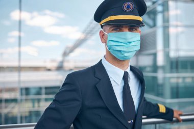 Tıbbi maskeli erkek pilot havaalanı terminalinde duruyor.
