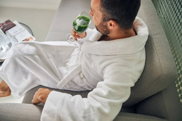 Vousatý mladý muž sedí na gauči a pije zelený smoothie — Stock fotografie
