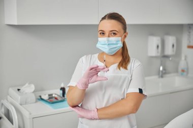 Tıbbi maskeli kadın kozmetik uzmanı elinde şırınga tutuyor.