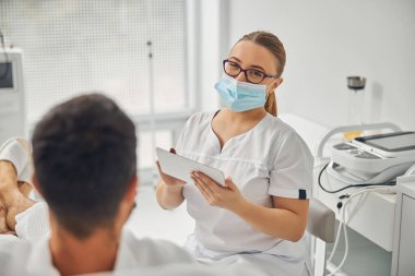 Tablet bilgisayarlı kadın estetisyen erkek hastayla konuşuyor.