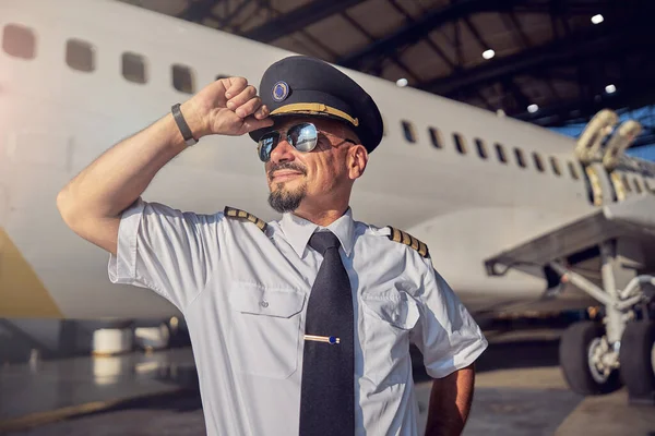 Hombre barbudo seguro sosteniendo el sombrero de pilotos en la cabeza mientras envía tiempo al aire libre — Foto de Stock