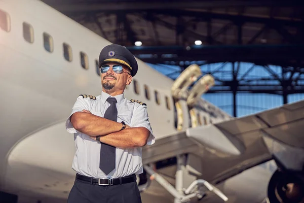 Przystojny, pewny siebie pilot w mundurze biznesowym i okularach przeciwsłonecznych ustawionych przed kamerą fotograficzną na lotnisku — Zdjęcie stockowe