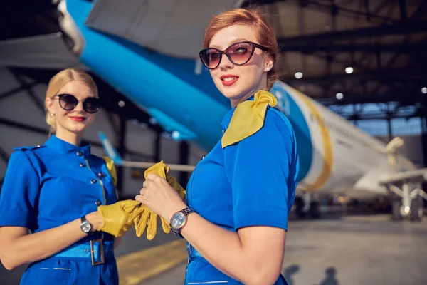 Šťastný úsměv dva letušky v uniformě a kožené rukavice pózující na kameru v leteckém hangáru — Stock fotografie