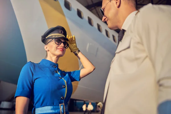 Charming woman flight attendant in hat and suit welcoming pilot near the airplane — Stock Photo, Image