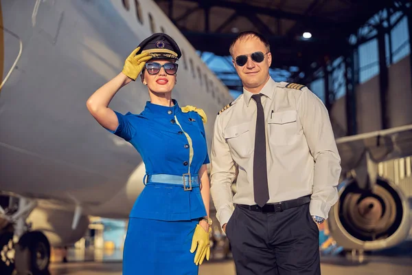 Elegancka stewardessa z kapitanem pozującym przed kamerą fotograficzną na lotnisku — Zdjęcie stockowe