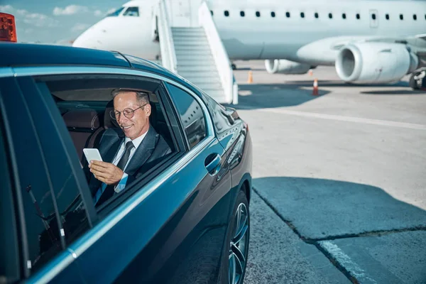 Cheerful businessman with smartphone in car at airport — Stock Photo, Image