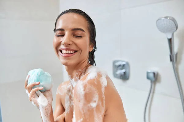 Χαμογελαστή νεαρή γυναίκα με αφρό στο σώμα της κρατώντας το μπάνιο loofah — Φωτογραφία Αρχείου