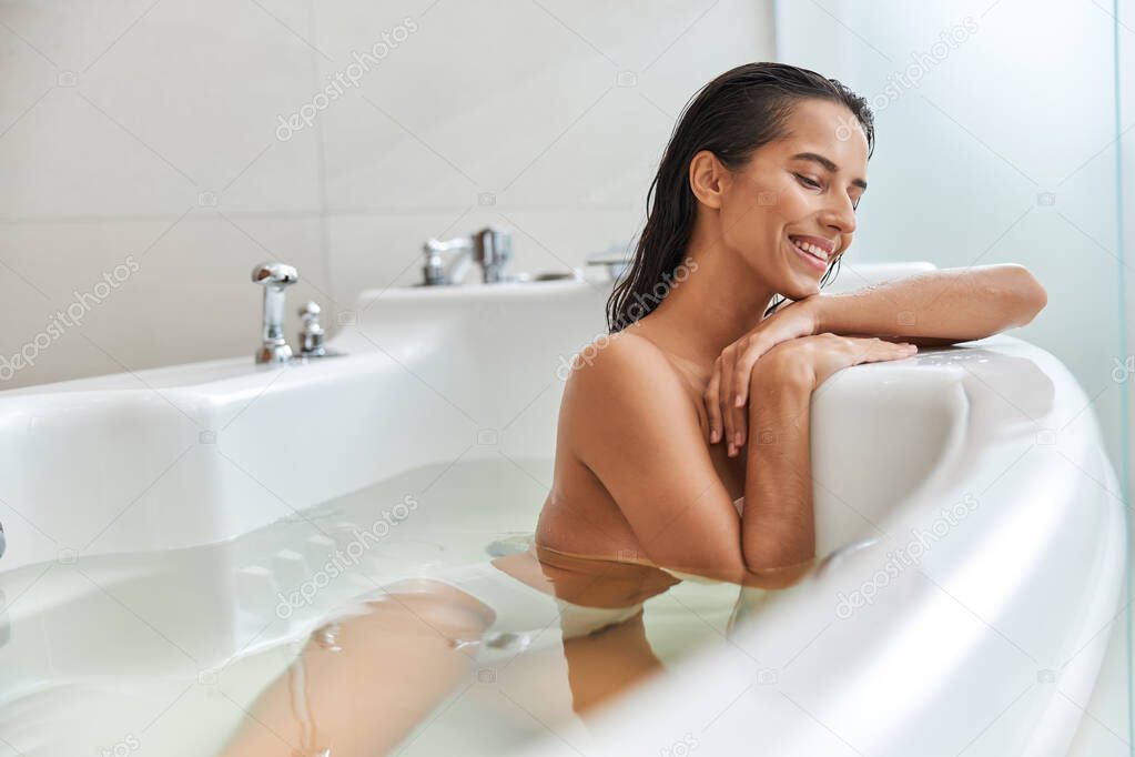 Charming young woman lying in white bathtub at home