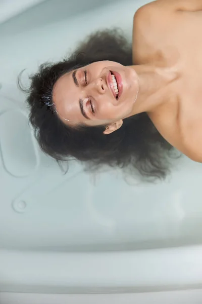 Cheerful young woman lying in water in bathtub