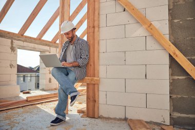 Sıkı şapkalı hırslı inşaat işçisi dizüstü bilgisayarını tutuyor.