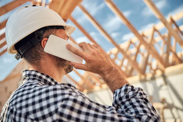 Construtor jovem de cabelos escuros em um capacete protetor fazendo um telefonema — Fotografia de Stock