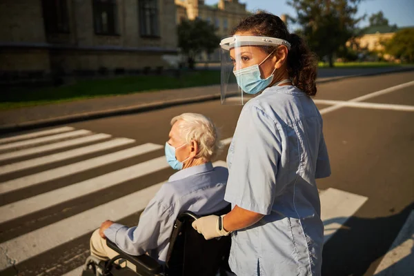 Vidám nő kék orvosi egyenruhában sétál egy idős férfival a városban. — Stock Fotó
