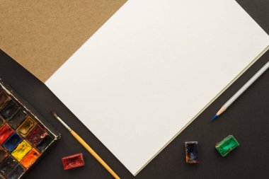 Sanatçının çalışma yeri: eskiz defterini aç, suluboya boya boya boya boya ve fırçalar koyu arkaplanda resim yapmak için. Hobi ve el işi konsepti. Üst görünüm.