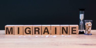 MIGRAIN kelime ahşap küpler üzerinde, küpler tahta ve gri arka planda durur, küplerin yanında haplar ve bir kum saati. Tıbbi ve sağlık hizmetleri kavramı