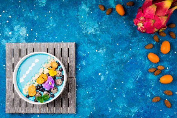 Blue smoothie bowl with fruits, berries, nuts and flowers. Tropical healthy smoothie dessert. Healthy food, vegetarian, diet concept. Top view. Copy space