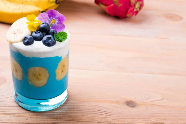 Tropical smoothie with banana fruits, blueberries and flowers on wooden background. Healthy smoothie dessert. Healthy food, vegetarian, diet concept. Copy space