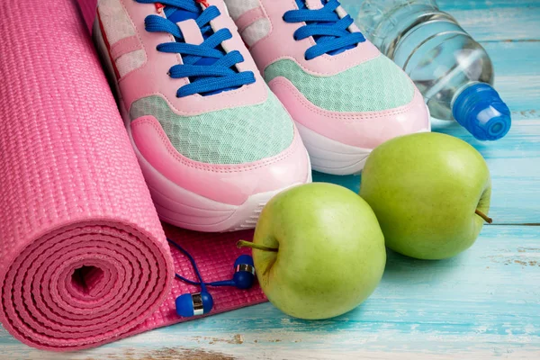 Pink yoga mat, sport shoes, bottle of water and apples on blue w