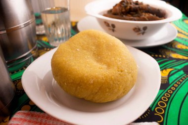 Beyaz bir tabağa yerleştirilmiş lezzetli sarı Garri veya Eba tepsisi renkli yeşil ve sarı Afrika desenli masa örtüsü üzerine kurulmuş yerel Nijerya sebze çorbası ile yenmeye hazır.