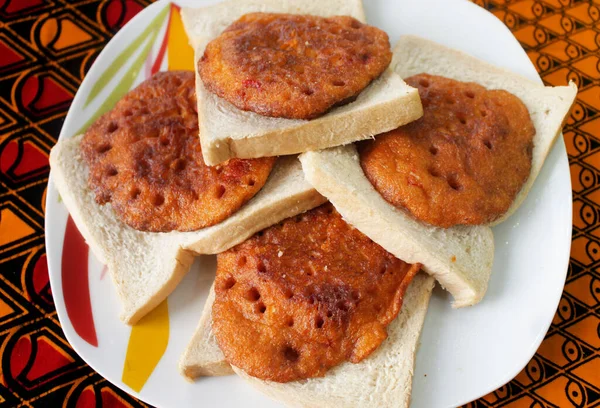 Dört parça ev yapımı fasulye gözlemesi ya da renkli Afrika kırmızı ve sarı desenli masa örtüsü üzerinde bir dilim ekmekle servis edilen Nijeryalı Akara.