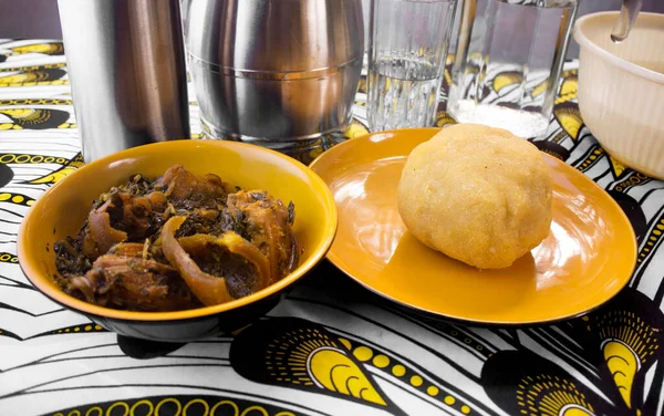 Lezzetli bir Nijerya Garri 'si, Eba' sı ve çeşitli et ve balıkların bulunduğu sebze çorbası turuncu renkli bir kâse ve tabakta renkli beyaz ve sarı Afrika desenli masa örtüsü üzerinde servis edilir.