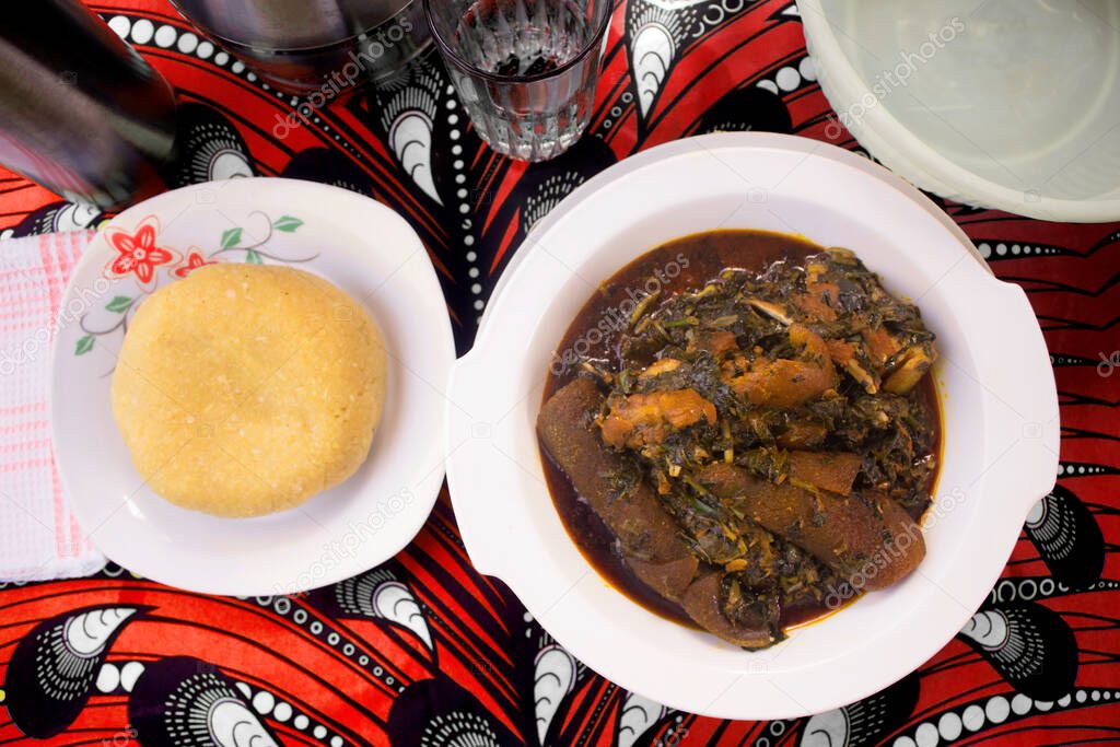 Top view of a delicious meal of Eba or Garri served with a bowl of Nigerian vegetable soup cooked with assorted meat, dried fish, cow skin (ponmo or kpomo) and set on a colorful red and white African pattern table cloth