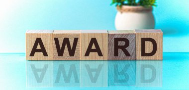 AWARD 'ın metni ahşap küplerde, mavi arkaplanda. Yazılı metin parlak yüzeyden yansıtılır. Arka planda saksıda bir çiçek var..