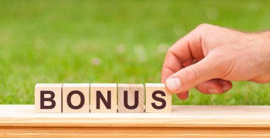 Elinde tahta küp bloğu olan bir adam yeşil çimenlik arka planda BONUS iş sözcüğüyle. İnsan eliyle BONUS 'u engelle. İkramiye işteki motivasyondur..
