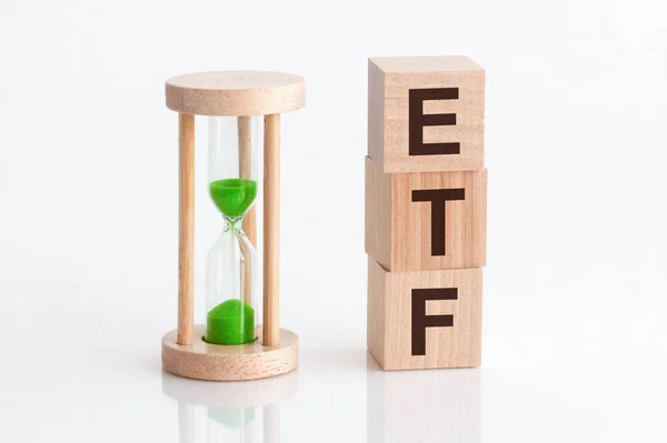 Close-up of an hourglass next to wooden blocks with the text ETF. ETF - exchange-traded fund - text in wooden building blocks, white backgrounds.