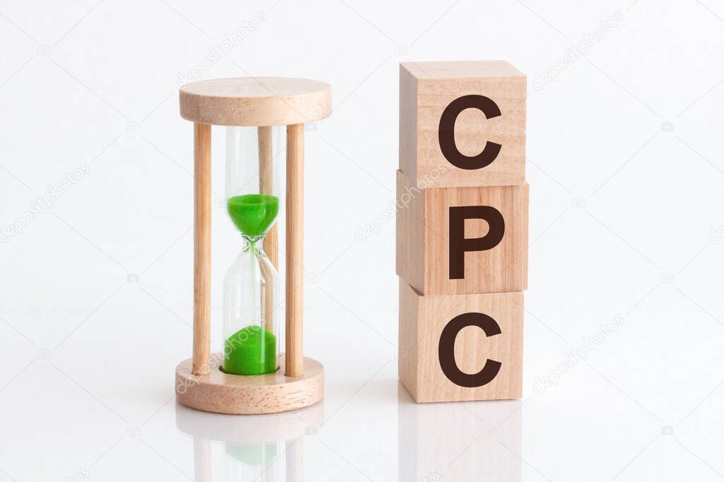 Close-up of an hourglass next to wooden blocks with the text CPC. CPC - Cost Per Click - text in wooden building blocks, white backgrounds.