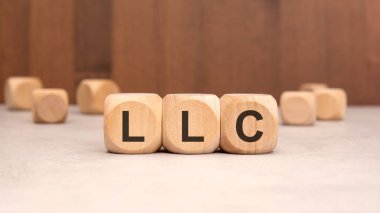 Tarafsız zemin üzerinde LLC yazılırken tahta bloklar bulanık arka plan, iş yapısını, sınırlı sorumluluk şirketini, girişimciliği, şirket oluşumunu ve yasal varlık kavramını sembolize ediyor.