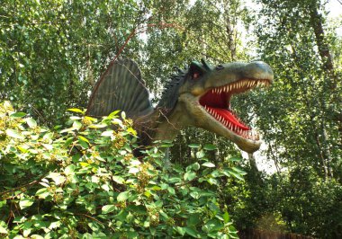 Açık çeneli büyük yeşil dinozor yapraklı parkta duruyor.