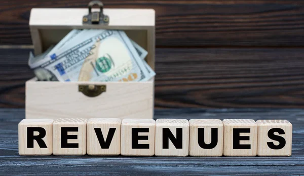 Revenues - word on cubes with a chest of money against a dark background. Business and finance concept