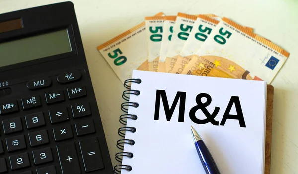 M&A (Mergers and Acquisitions) word in a notebook against the background of calculitar and banknotes. Business and finance concept.