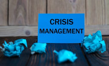 CRISIS MANAGEMENT - Buruşuk kağıt parçaları ile koyu ahşap zemin üzerinde mavi kağıt üzerinde kelimeler. İş ve finans kavramı.