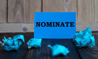 NOMINATE - mavi kağıda yazılmış kelimeler koyu ahşap arka planda ve buruşuk kağıt parçaları. İş ve finans kavramı.