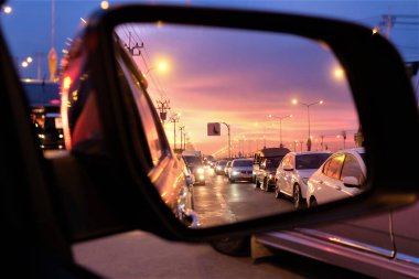 Arabanın dış aynasında seçici bir bakış. Birçok arabanın akşam vakti kırmızı-turuncu gökyüzünün altında yağmur yağar gibi trafik sıkışıklığı vardır..