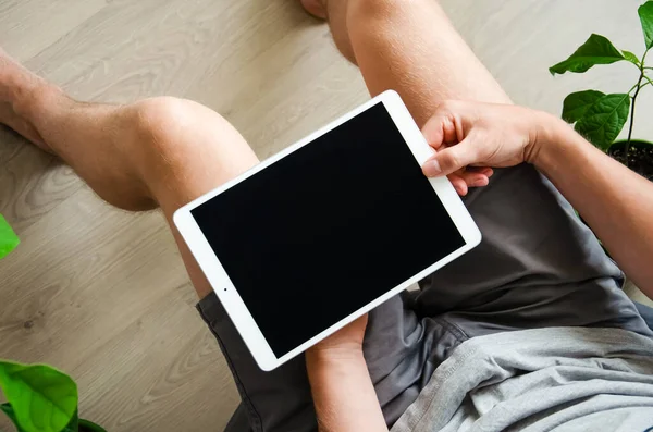 Top view of a tablet on a caucasian man\'s lap. Man uses a laptop while sitting on the floor of a house near home flowers. Working home concept. Using tablet for education and for work.