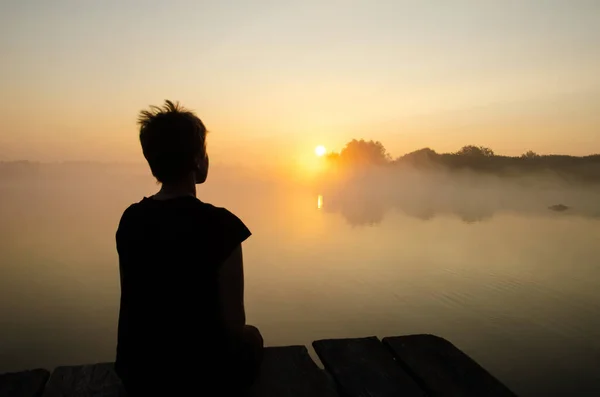 Silhouette of a girl sitting on the pier against the sunset sky with copy space. The girl looking at the rising sun over the lake in the fog. Reflection of the sun in the water surface.