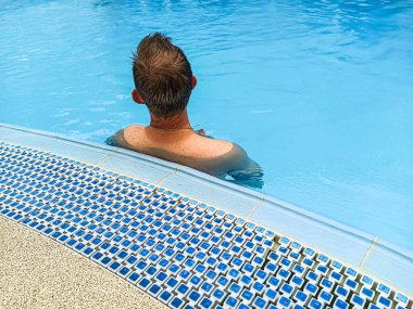 Kafkasyalı genç bir adam sırtı temiz su dolu bir havuzda duruyor. Mavi dekoru ve mavi suyu olan bir havuzda genç bir adamın arkasında. Havuzda yaz tatili. Yaz günü havuzda serinleyen adam..