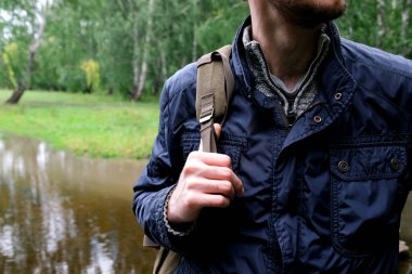 Mavi ceketli bir adam ormanda sırt çantalı duruyor. Bir dere ve yeşil ağaçlar görülebilir. Yüzü yok.