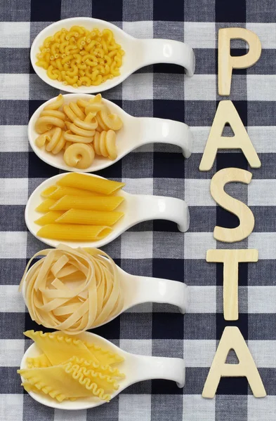 Pasta written with wooden letters on checkered cloth and uncooked pasta on porcelain spoons