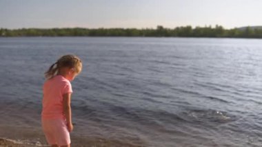 Küçük kız nehrin kenarında durur ve suya taş atar.