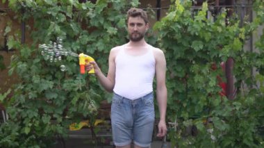 Sakallı, çocuksu bir genç adam ayakta duruyor ve oyuncak bir baloncuktan yan tarafa sabun köpükleri fırlatıyor.