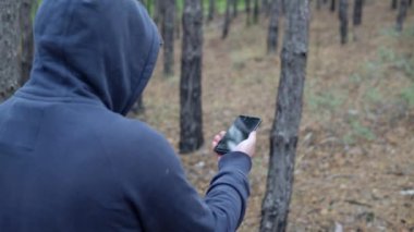Kapüşonlu bir adam akıllı telefonuyla ormanda yürüyor.