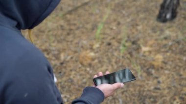Kapüşonlu bir adam akıllı telefonuyla ormanda yürüyor.