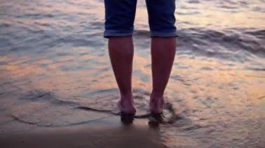 Bir adam kumlu bir sahilde duruyor ve dalgalar ayaklarını ıslatıyor.