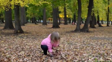 Parktaki neşeli bir kız düşmüş sarı yaprakları toplar ve fırlatır.